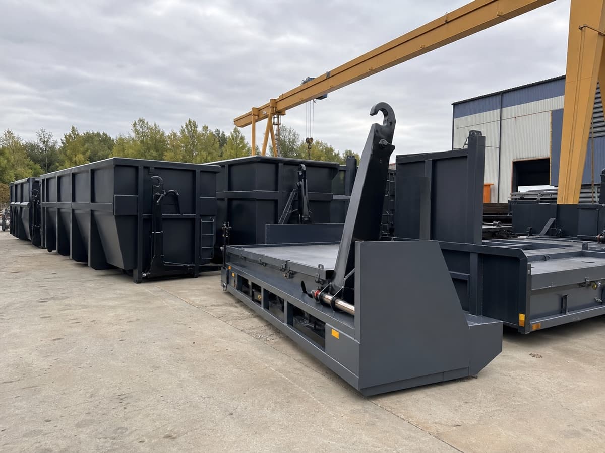 Row of newly painted welded steel hooklift containers and roll-off platforms outside a fabrication shop